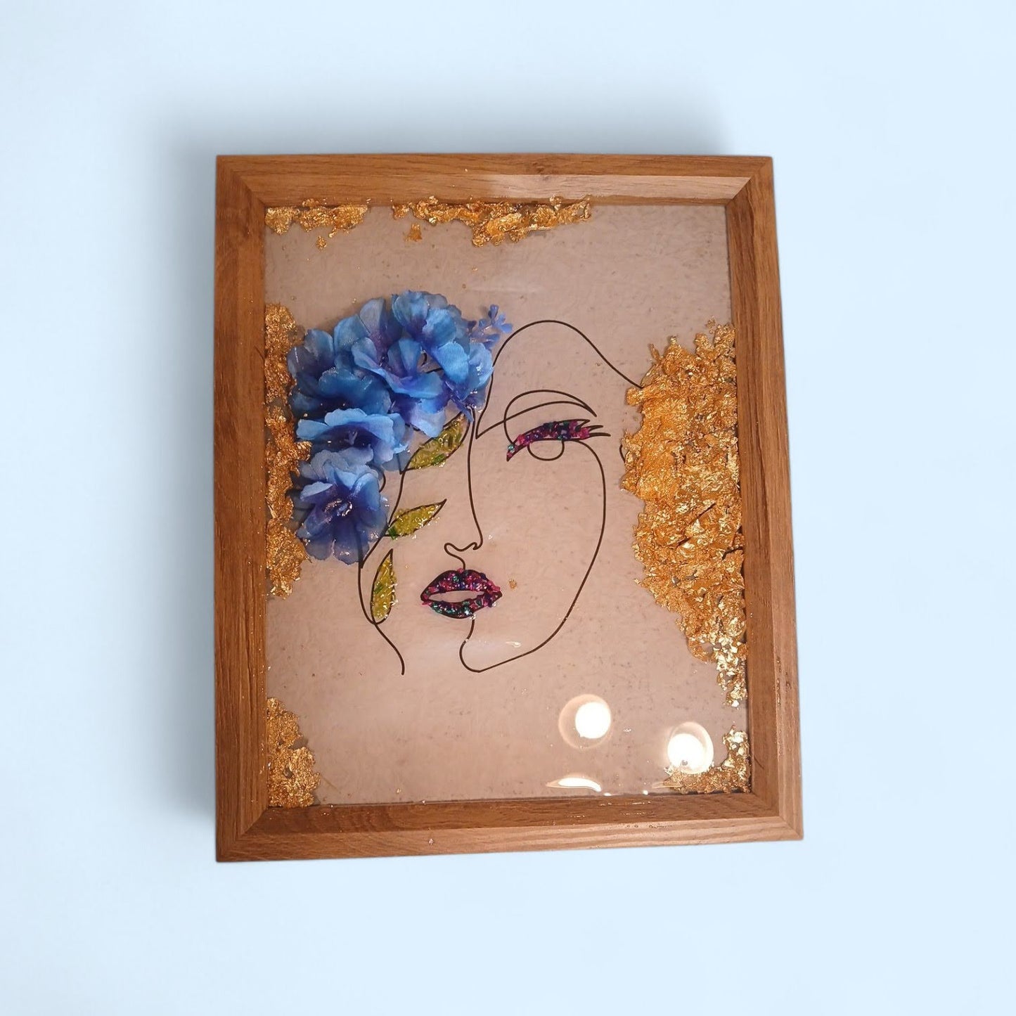Artistic depiction of a face with blue flowers and gold accents in a wooden frame.