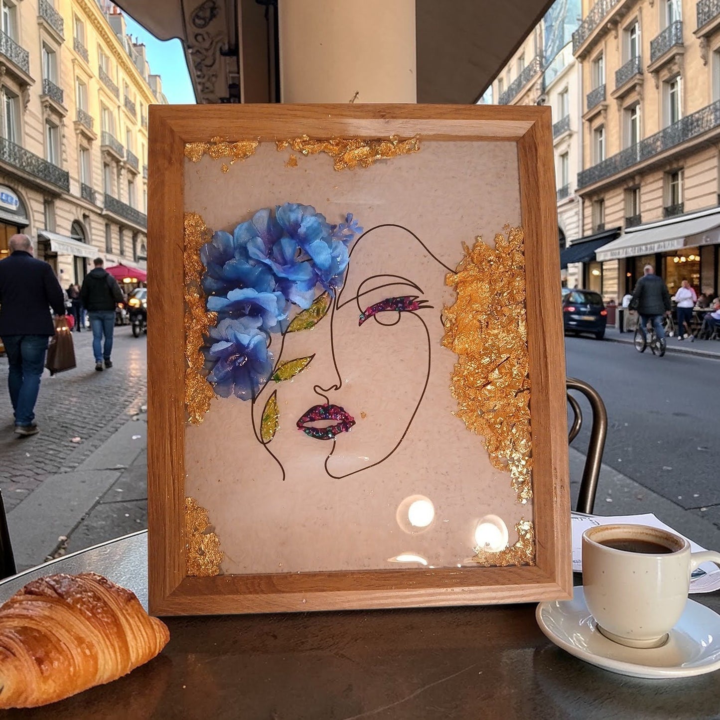 Framed artwork of a face with flowers and gold accents on a city street.