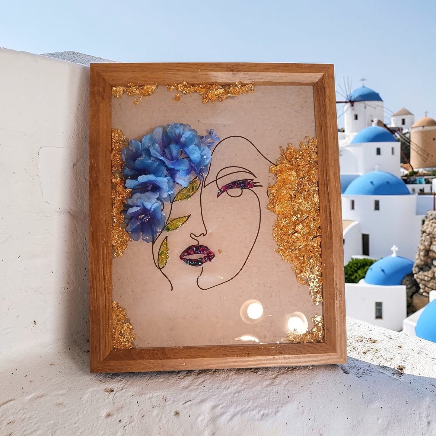 Artistic depiction of a face with flowers and gold accents in a wooden frame, set against a scenic background.