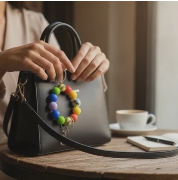 Person holding a gray handbag with colorful pom-poms on a wooden table.