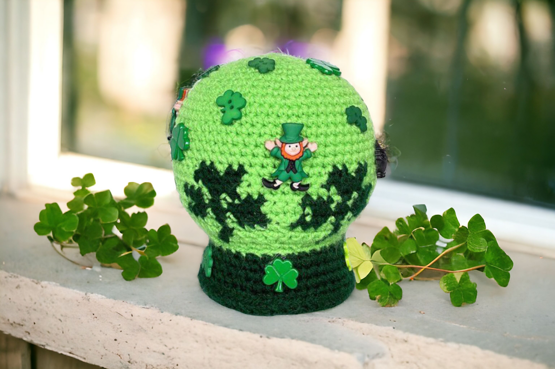 Green crochet sphere with shamrock patterns and a small figure on a wooden surface with leaves.
